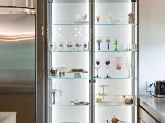 Showcasing a modern kitchen with a sleek, illuminated glass display cabinet filled with elegant glassware and decorative items.