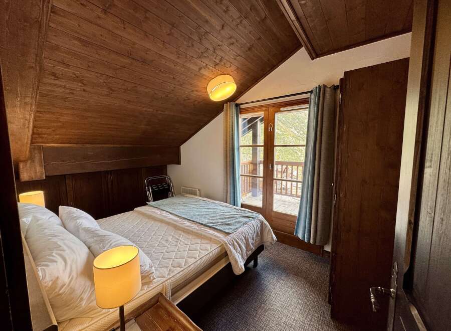 Experience cozy mountain living in this charming bedroom featuring a comfortable bed, warm wood-paneled walls, and a private balcony accessed through French doors.