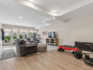 This versatile living space features a comfortable grey sofa, a modern desk setup with ergonomic chairs, and a dedicated home gym area complete with a treadmill and weights.