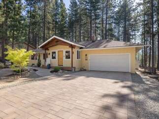 Charming single-story home nestled among tall pine trees, featuring a welcoming yellow exterior and a spacious paver driveway.