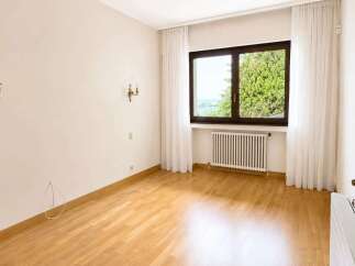 This empty room features a large window with white curtains, offering a glimpse of lush greenery and a distant body of water.