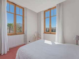 Experience natural light and scenic views from this charming bedroom, featuring two large wooden-framed windows that open up to a bright blue sky and lush greenery.
