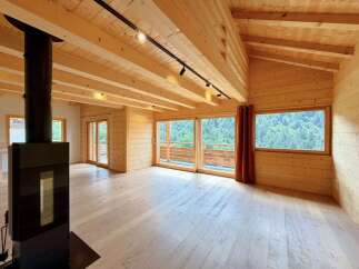 Experience modern mountain living in this stunning wooden interior featuring a sleek black wood-burning stove and expansive windows that frame breathtaking forest views.