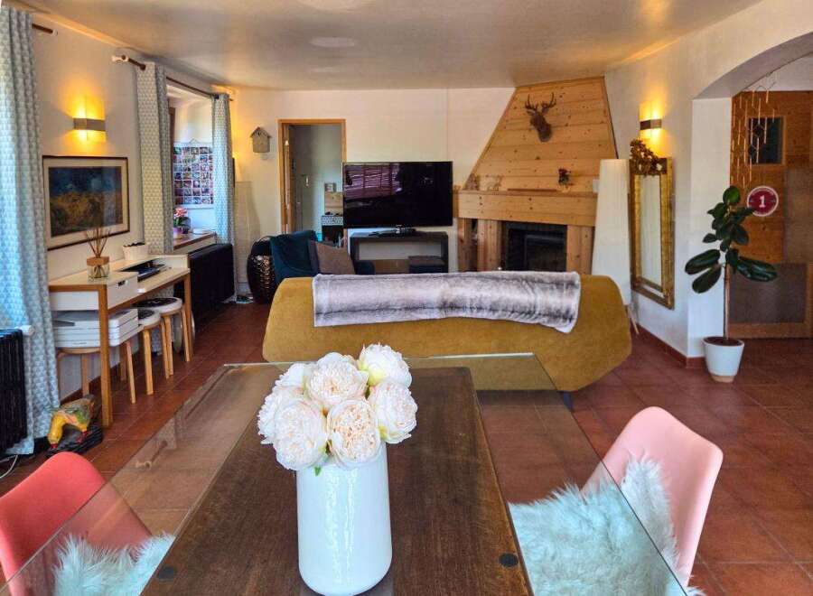 This cozy living room features a rustic wooden fireplace with a mounted deer head, a comfortable mustard-yellow sofa with a plush throw, and a modern desk area with stools.