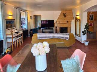 This cozy living room features a rustic wooden fireplace with a mounted deer head, a comfortable mustard-yellow sofa with a plush throw, and a modern desk area with stools.