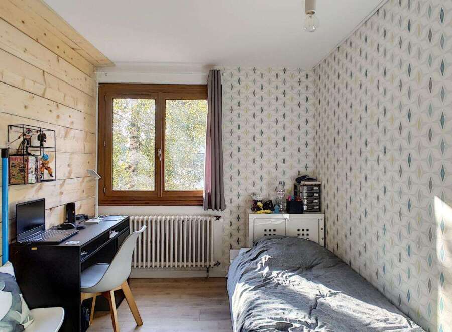 Discover this charming room featuring a cozy bed with grey bedding, a functional desk area with a laptop, and a large window offering natural light.