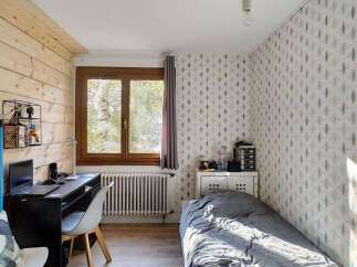 Discover this charming room featuring a cozy bed with grey bedding, a functional desk area with a laptop, and a large window offering natural light.