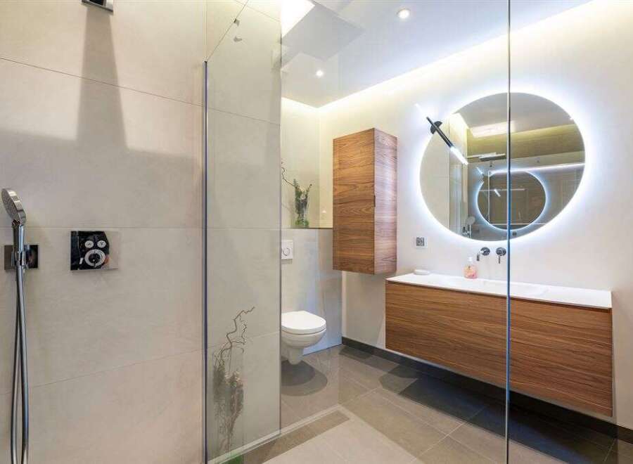 Experience modern luxury in this stunning bathroom featuring a sleek walk-in shower with a rainfall showerhead and a contemporary vanity with a backlit circular mirror.