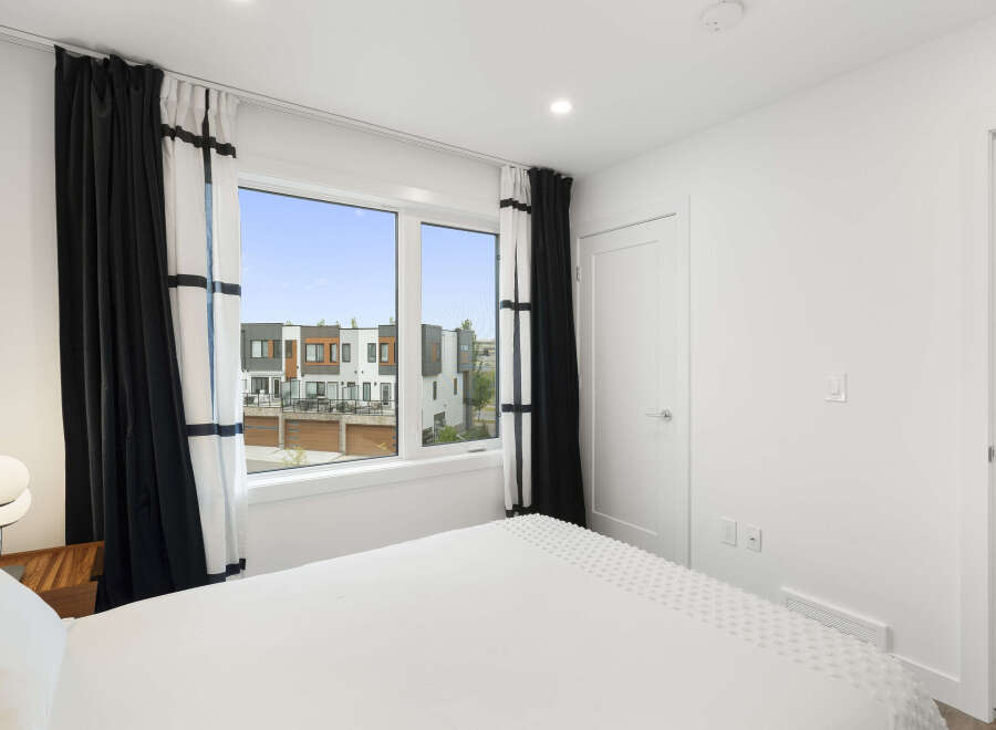 Experience modern living in this bright bedroom featuring a large window with scenic views of contemporary townhouses.