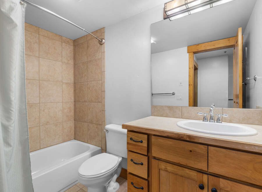 This bathroom features a tiled shower-tub combination with a white shower curtain, a white toilet, and a wooden vanity with a beige countertop and a white sink.
