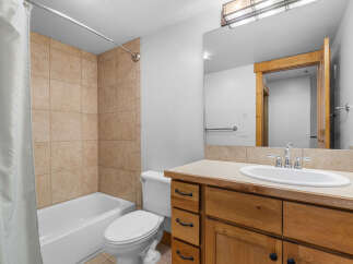 This bathroom features a tiled shower-tub combination with a white shower curtain, a white toilet, and a wooden vanity with a beige countertop and a white sink.