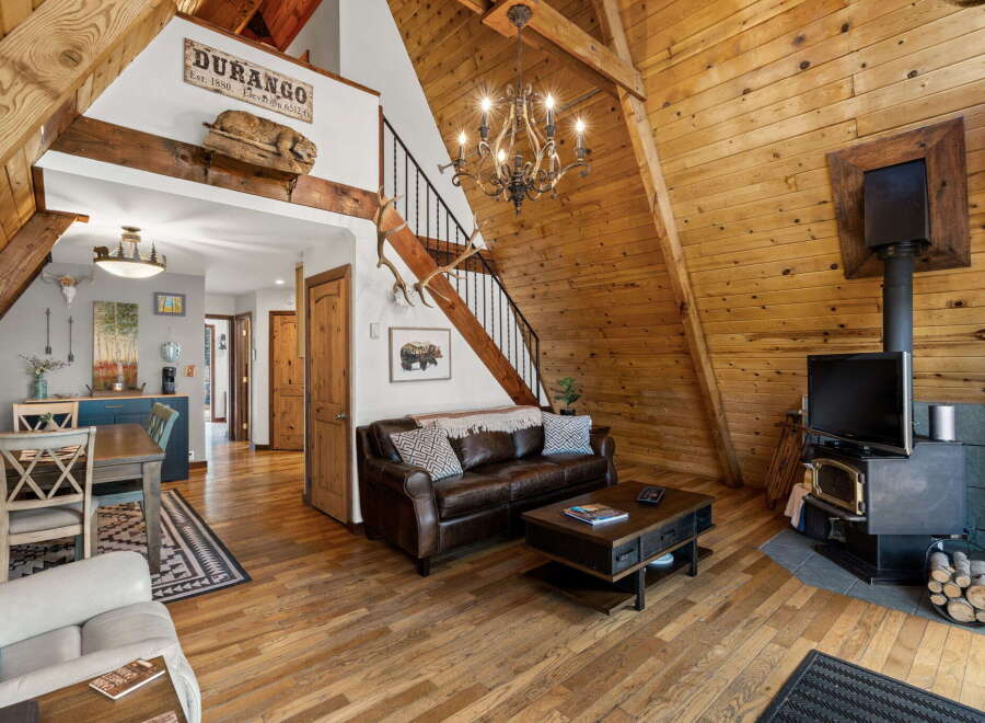 Experience rustic charm in this A-frame cabin living room, featuring a rich leather sofa, a striking chandelier, and a cozy wood-burning stove.