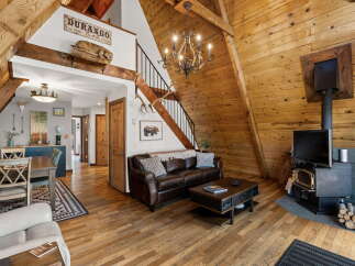 Experience rustic charm in this A-frame cabin living room, featuring a rich leather sofa, a striking chandelier, and a cozy wood-burning stove.