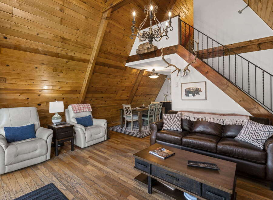 Experience rustic charm in this A-frame cabin living room, featuring warm wood paneling throughout and a cozy seating area with plush recliners and a leather sofa.