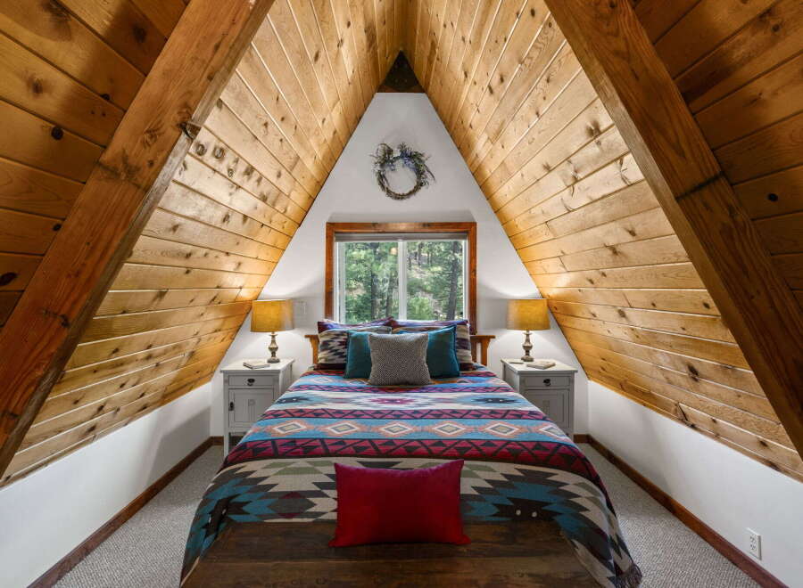 Experience rustic charm in this A-frame bedroom, featuring a cozy bed adorned with a vibrant, geometric-patterned comforter and plush pillows.