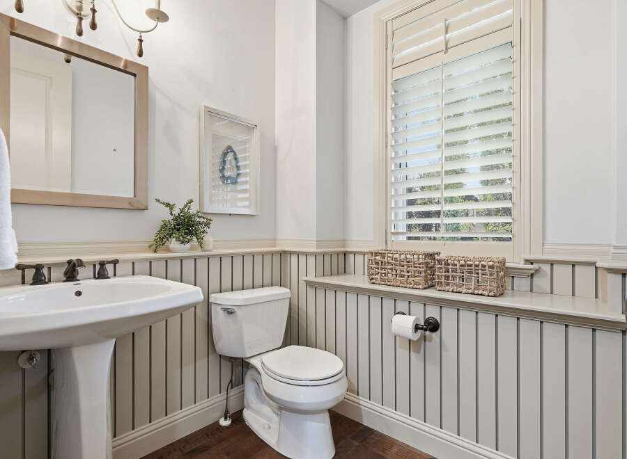 This charming bathroom features a classic pedestal sink with elegant bronze fixtures, complemented by a white beadboard wainscoting and dark wood flooring.