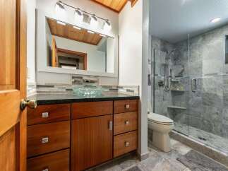 This modern bathroom features a sleek vanity with a glass vessel sink and ample storage, complemented by a spacious walk-in shower with elegant stone tilework.