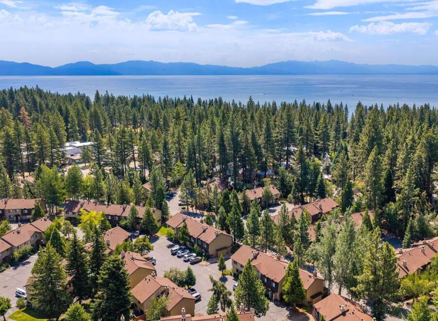 Discover this stunning aerial view of a residential community nestled amongst tall pine trees, offering a serene escape with a picturesque lake and distant mountains as a backdrop.