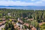 Discover this stunning aerial view of a residential community nestled amongst tall pine trees, offering a serene escape with a picturesque lake and distant mountains as a backdrop.