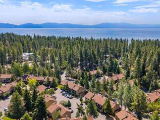 Discover this stunning aerial view of a residential community nestled amongst tall pine trees, offering a serene escape with a picturesque lake and distant mountains as a backdrop.