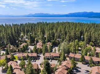 Discover this stunning aerial view showcasing a charming residential community nestled amidst a dense forest, with a breathtaking lake and distant mountains forming a picturesque backdrop.