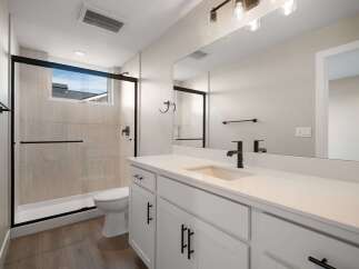 This modern bathroom features a sleek white vanity with black hardware, complemented by a spacious walk-in shower with a glass enclosure and light-toned tile.