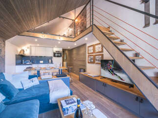 Experience modern alpine living in this stylish loft apartment, featuring a sleek kitchen, a cozy living area with a plush blue sofa, and a unique mezzanine accessible by a wooden staircase.