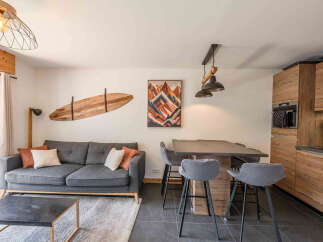 This modern living and dining area features a comfortable grey sofa with accent pillows, a stylish wooden surfboard wall art, and a sleek dining table with bar stools.