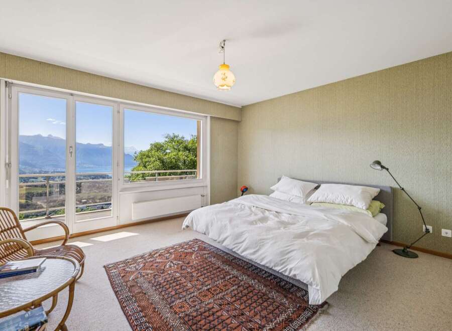 Experience breathtaking panoramic views from this serene bedroom, featuring a large window overlooking a stunning lake and mountains.