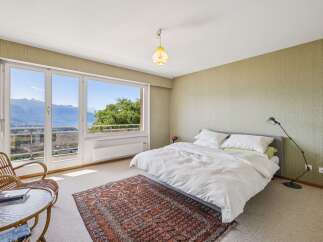 Experience breathtaking panoramic views from this serene bedroom, featuring a large window overlooking a stunning lake and mountains.