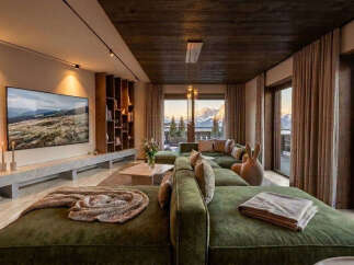 Experience luxury mountain living in this stunning living room, featuring a plush green sectional sofa and a large flat-screen TV displaying a scenic landscape.