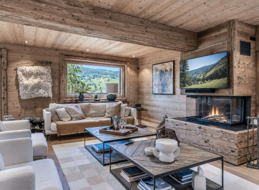 Experience luxurious mountain living in this stunning chalet, featuring a cozy living room with a modern fireplace and a large window offering breathtaking panoramic views of the rolling hills and lush greenery.