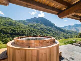 Experience ultimate relaxation with this stunning outdoor wooden hot tub, offering breathtaking panoramic views of lush green mountains and a clear blue sky.