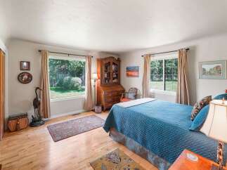 This charming bedroom features a large blue quilted bed, a vintage wooden secretary desk, and two large windows offering serene views of the lush backyard.
