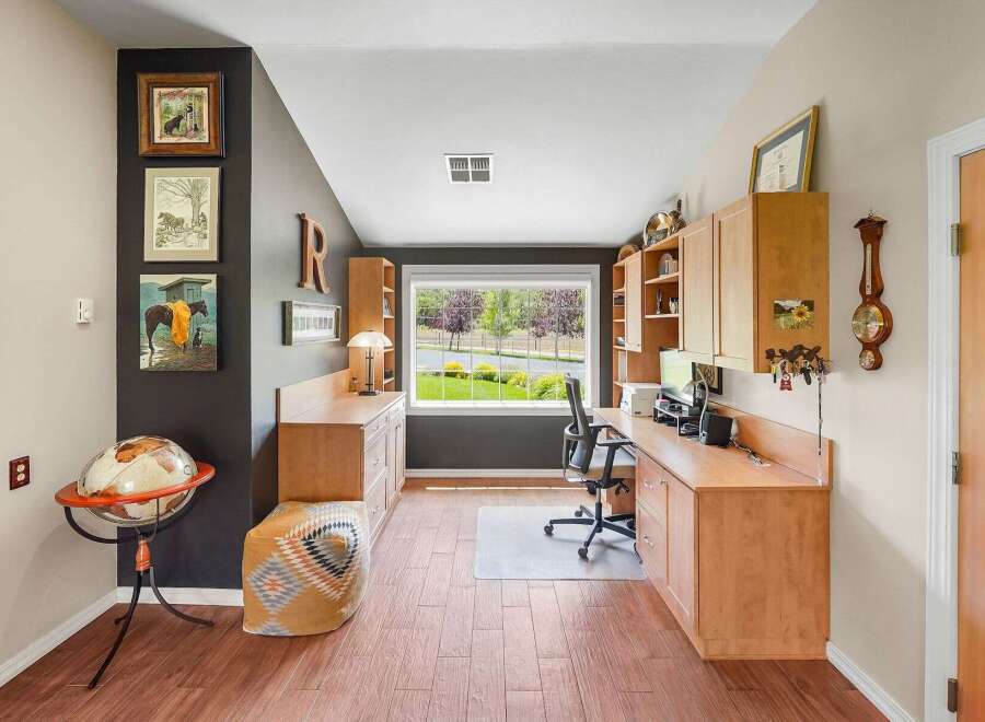 This home office features a spacious built-in desk with ample storage and a large window overlooking a lush green yard.