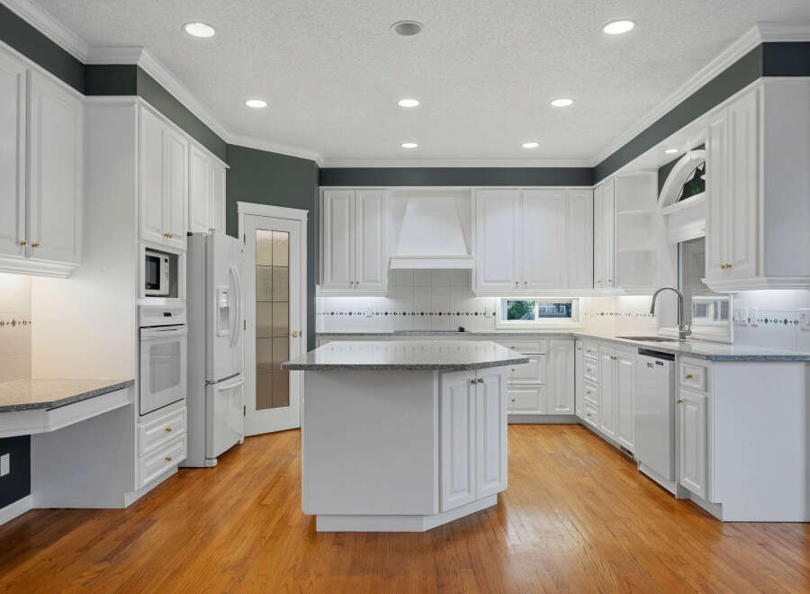 This spacious kitchen boasts pristine white cabinetry, complemented by a large central island with a granite countertop, perfect for meal preparation and casual dining.