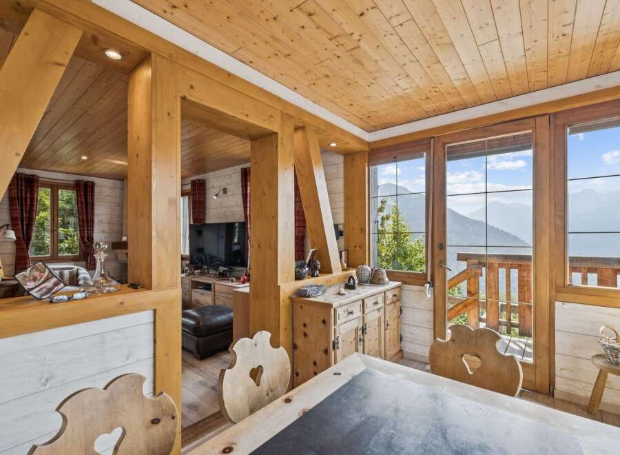 Experience breathtaking mountain views from this charming wooden chalet, featuring a cozy dining area with rustic wooden chairs and a large window overlooking the stunning landscape.