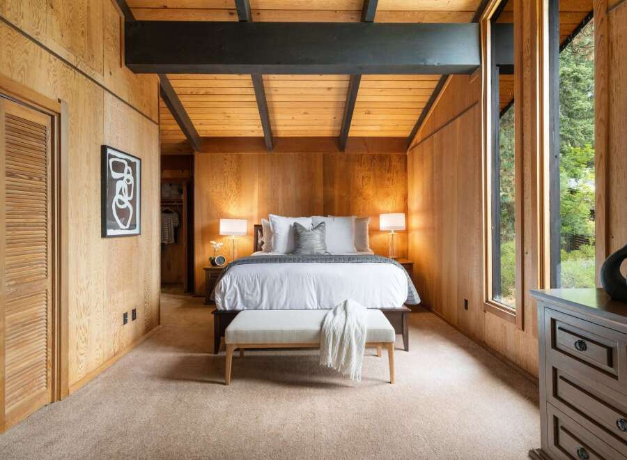 Experience rustic charm in this beautifully appointed bedroom, featuring warm wood-paneled walls and a vaulted ceiling with exposed beams.