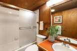 This charming bathroom features a classic white tiled shower/tub combo with a frosted glass sliding door, complemented by warm wood paneling on the walls and ceiling.