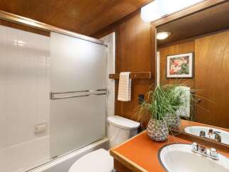 This charming bathroom features a classic white tiled shower/tub combo with a frosted glass sliding door, complemented by warm wood paneling on the walls and ceiling.