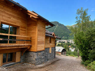 Experience mountain living in this stunning wooden chalet, featuring a stone foundation and a spacious balcony overlooking a picturesque village nestled in the valley.