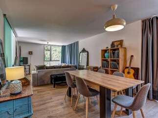 This spacious living and dining area features a comfortable grey sectional sofa, a large wooden dining table with upholstered chairs, and a stylish wooden bookshelf filled with decor and books.