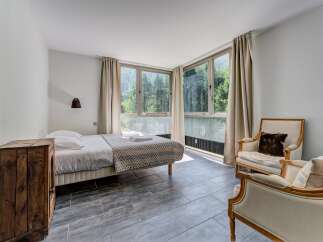 Experience serene mountain living in this charming bedroom featuring a comfortable bed, a rustic wooden dresser, and large windows offering breathtaking forest views.