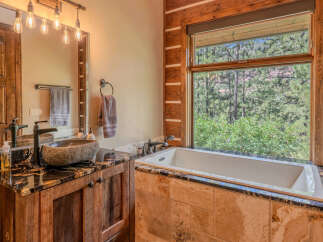 Experience rustic luxury in this stunning bathroom, featuring a unique stone vessel sink atop a granite countertop and a deep soaking tub with a breathtaking forest view.