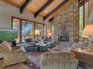 Experience breathtaking lake and mountain views from this stunning living room, featuring a grand stone fireplace and vaulted wood-beamed ceilings.