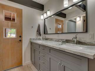 This modern bathroom features a sleek double vanity with a granite countertop and gray cabinetry, complemented by a large, illuminated mirror.