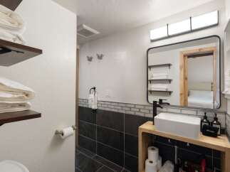 This modern bathroom features a sleek vessel sink atop a rustic wooden vanity, complemented by a large black-framed mirror reflecting a neatly organized shelving unit.