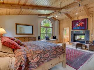 Experience rustic elegance in this charming bedroom, featuring a cozy fireplace and a large arched window offering serene forest views.