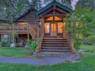 Charming log cabin nestled amongst tall trees, featuring a welcoming wooden porch with a stone pathway leading to the entrance.