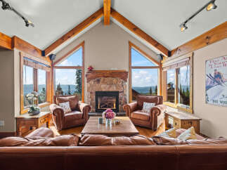 Experience breathtaking mountain views from this stunning living room, featuring a grand stone fireplace and vaulted wooden beam ceilings.